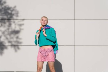 Yeşil tişörtlü ve pembe şortlu genç bir Latin adam omuzlarında gurur bayrağı olan bir fotoğraf için poz veriyor, öğleden sonra güneşinin altında kameraya bakıyor..