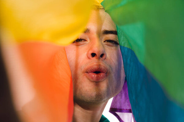 Close-up front view of Latin Gay Man looking at camera blowing a kiss covered by a pride flag. Concept of humor and lightheartedness, as the man is making a silly gesture while holding the colorful flag