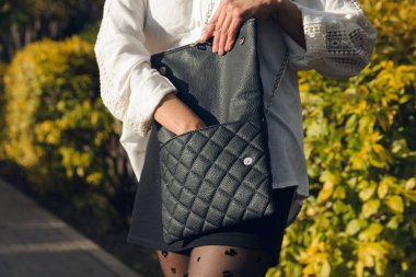 Elini deri çantasının içine sokup, parkta telefonunu arayan tanınmayan bir kadının beline yakın çekim. O, beyaz bir gömlek ve siyah kısa pantolon giyiyor.