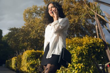 Genç bir Latin kadın arka planda ağaçlarla gün batımında parkta dikilirken cep telefonuyla konuşuyor. O, beyaz bir gömlek ve siyah kısa pantolon giyiyor, telefonda dikkatle dinliyor.