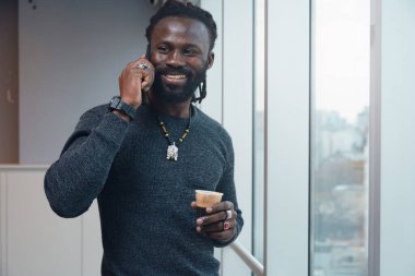 Bir Mutlu Adam elinde bir fincan kahve tutarken cep telefonuyla konuşuyor. Gülümsüyor ve birlikte çalışırken pencerenin kenarında durarak eğleniyor..