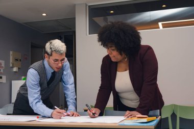 İki genç kadın ve erkek, bir proje üzerinde birlikte çalışıyorlar. Konuşuyorlar ve bir kağıda not alıyorlar. Kalemlerle ve kitaplarla dolu bir masaya yazı yazıyorlar, kurumsal ve zarif bir görünüşleri var..