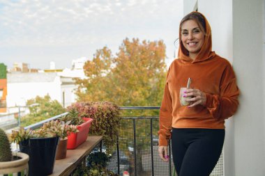 Evinin balkonundaki genç Latin kadın bitkilerinin yanında sırıtarak duvara yaslanıyor. Kameraya bakıyor, elinde bir eş içeceği, arka planda fotokopi alanı olan bir gökyüzü tutuyor..