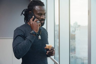 Bir adam elinde kahve tutarken cep telefonuyla konuşuyor. Kolye ve yüzük takıyor.