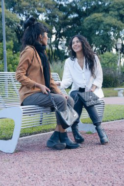 İki Latin kadının öğleden sonra parkta keyif çattığı sosyal ağlar için dikey görüntü. Biri kahverengi ceketli, diğeri beyaz gömlekli.