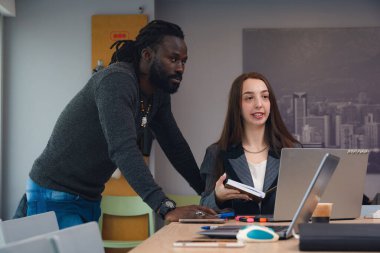Yetişkin bir adam ve kadın ofiste birlikte çalışırken beyaz tahtaya doğru bakıyor ve dizüstü bilgisayarla çalışan kadına bir şeyler öğretiyor..