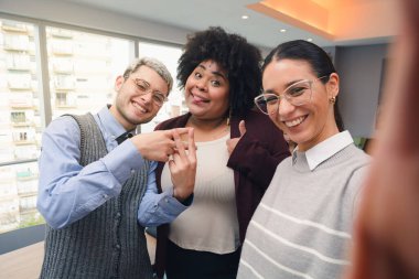 Ofiste iki genç kadın ve bir adam gülümsüyor ve özçekim fotoğrafı için poz veriyorlar. Resmi kıyafetleri var ve telefonu gözlüklü bir kız tutuyor, perspektif de telefonun kamerasından.