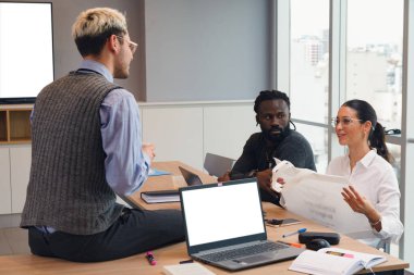 Masanın üstündeki dizüstü bilgisayarın yanında oturan genç işadamı, tasarımcı işbirlikçilerinin talimatlarına dikkat ederek, onlara işin nasıl yapıldığını gösteriyor..