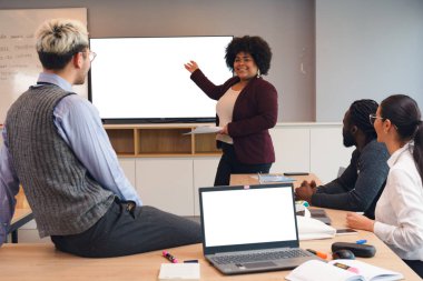 Beyaz perdenin önünde duran Afro Latin Kadın iş arkadaşları ve ortaklarına farklı konuları sunuyor. Meslektaşları onu dinliyor ve izliyorlar.