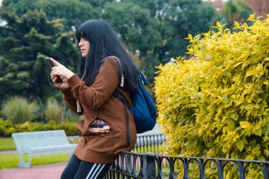 Genç Latin Kadın sakince park yerinde sosyal medyayı kontrol ediyor, çalıların önünde korkuluklara oturuyor, kahverengi kazak ve sırt çantası giyiyor, teknoloji kavramı, kopyalama alanı.