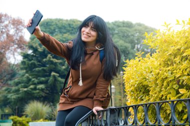 Parkta oturan genç Latin kadın cep telefonuyla cep telefonuyla fotoğraf göndermekle meşgul. Kahverengi süveter giyiyor, çalılıkların yanındaki korkuluklara oturuyor, fotokopi çekiyor.