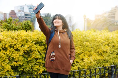 Mutlu genç Latin Kadın sakince park yerinde duruyor. Telefonla kendi fotoğrafını gönderiyor. Arka planda çalılar ve gökyüzü, teknoloji ve sosyal ağlar kavramı var..