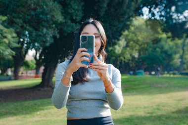 Parktaki genç Latin kadın gülümsüyor ve fotoğraf çekerken yüzünü cep telefonuyla kapatıyor, cep telefonunu yüzünün önünde, ön görünüşünün önünde, teknoloji kavramının önünde tutuyor.