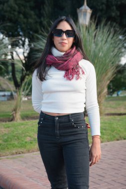 Genç Latin kadının sosyal ağları için dikey portresi. Parkta iyi davranışlı, güneş gözlüğü ve eşarpla poz veriyor..