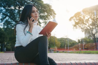 Park basamaklarında oturan genç kadın molada kitap okurken telefondaki sesli mesajı dinliyor..