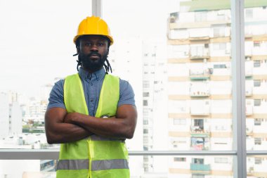 İnşaat ve bakım sektöründe çalışan Afrikalı bir işçinin portresi. Kollarını kavuşturup kameraya bakıyor. Şal, güvenlik kaskı ve mavi kıyafetler giyiyor.