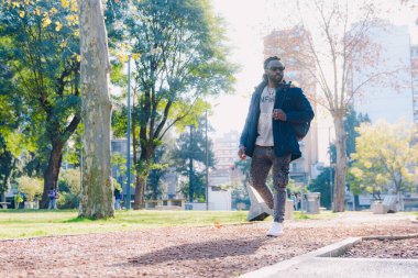 Afrika kökenli yetişkin bir adam, sırt çantası ve güneş gözlükleriyle, parka geliyor, yürüyor, etrafa bakıyor, fiziksel egzersiz alanı arıyor..