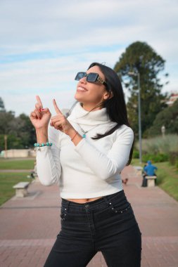 Güzel görünümlü, hoş tavırlı, günlük kıyafetlerle gülümseyen genç bir kadın parkta dikey görüntüsüyle gökyüzünü işaret ediyor.