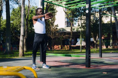 Rastalı ve sakallı siyah adam kolsuz gömlek ve siyah eşofman altı giyiyor. Parkta durmuş kollarını ve omuzlarını uzatarak antrenman yapmaya hazırlanıyor.