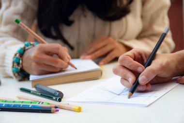 Evde çizim yapmak, tanınmayan iki kişiyi kapatmak, kağıt üzerinde çizim yapan kadın elleri, masada renkli kalemlerle oturmak, kopyalama alanı, sanat konsepti..