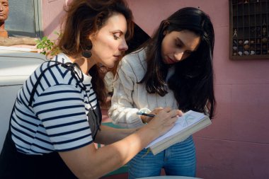 Bayan öğretmen çarşafta düzeltmeleri evdeki resim atölyesinde öğrencisine gösteriyor. Öğrencisi onu dikkatle izlerken o da çalışma kağıdını tutuyor ve üzerine yazıyor.
