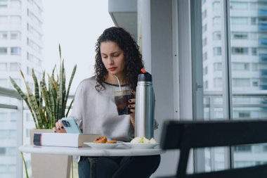 Girişimci, kıvırcık saçlı Latin Kadın cep telefonuyla oteldeki interneti tarıyor, içki içiyor ve kahvaltı yapıyor. Masanın üzerinde bir kitap var..