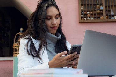 Genç Latin Girişimci Kadın evde cep telefonundaki mesajlara bakarak mutlu bir şekilde çalışıyor, dijital işlerini yönetiyor, e-posta mesajlarını cevaplıyor ve uygulama bildirimlerini kontrol ediyor.