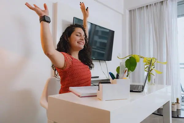 stock image Young Venezuelan woman at home with laptop sitting with arms raised celebrating her financial gains, she is smiling and looking at the camera happily after checking the reports in the digital wallets.