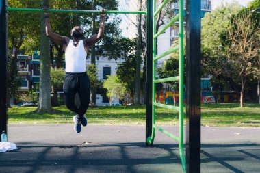 Beyaz tişörtlü güçlü siyah adam, parkta barfiks çekiyor. Rasta ve sakal takıyor. Kas kütlesi oluşturmak için beden eğitimi alıyor..