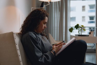 Oturma odasındaki kanepede oturan genç Latin kadının yan görüntüsü cep telefonuyla mesajları kontrol ederken arka planda bir pencere var.