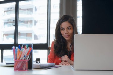 Kırmızı takım elbiseli Venezüellalı girişimci kadın ofiste dizüstü bilgisayarıyla oturmuş telefonuyla kendini kaydediyor, ortak çalışma alanında sosyal ağlarda canlı yayın yapıyor..