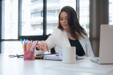 Beyaz elbiseli genç Latin kadın ofiste dizüstü bilgisayarla çalışıyor, telefonu masadan alıyor, iş konsepti, kopyalama alanı..