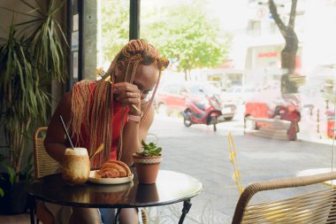 Genç Afrikalı bir kadın bir kafede kahvaltı yapıyor ve o kadar çok güldüğü için yemek yiyemiyor. Kolunu masaya dayayıp gülüyor. Masada kahve ve kruvasan var.