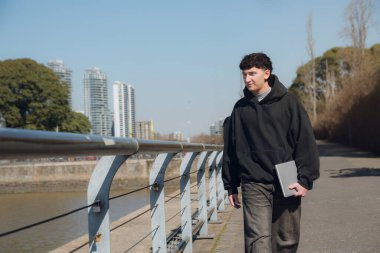 Genç erkek içerikli yaratıcı dizüstü bilgisayarıyla şehirde yürüyor, kalın bir süveter giyiyor ve yan tarafa bakıyor, arka planda bir bina ve fotokopi alanı olan açık bir gökyüzü var.