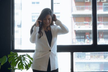 Ofiste arkasında pencere olan genç Latin bir kadın telefonla konuşuyor, eli başının üstünde gülümsüyor, beyaz ceket giyiyor, teknoloji konsepti kopyalama alanı kullanıyor.