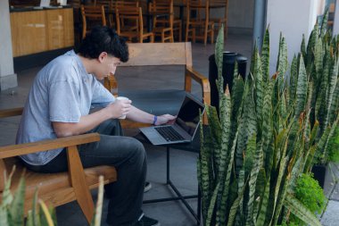 Genç Arjantinli bir adam bir kafede oturuyor, laptopuyla çalışıyor, kahve içiyor ve dinleniyor, ön planda bitkiler var, ahşap ve deri bir koltukta oturuyor, kopyalama alanı.