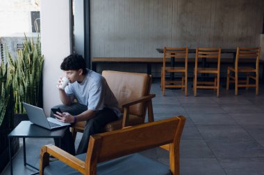 Arjantinli genç girişimci kahve içerken ve kafede dizüstü bilgisayarla çalışırken telefona bakıyor, ahşap ve deri koltukta oturuyor, arkasında fotokopi alanı olan gri bir duvar var.