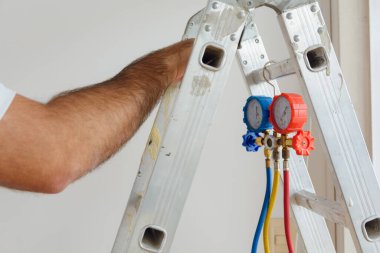 Katlanan alüminyum merdivenden sarkan basınç göstergesi ve tanınmayan bir adamın erkek eli kapalı kapılar ardındaki merdivenin gidonuna dayanıyor..