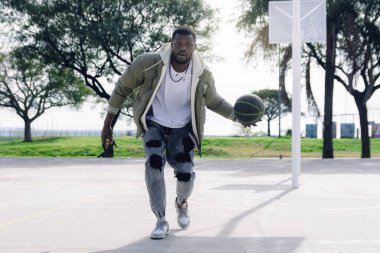 Günlük giysiler içinde, basketbol sahasında kameranın önünde top sallanırken kendini eğlendiren genç Afrikalı bir adam zorlu bir yüzle kameraya bakıyor..