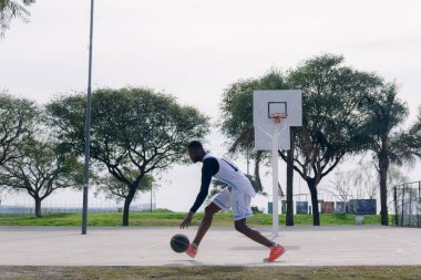 Beyazlar içinde Afrikalı etnik kökenli genç bir adam basketbol sahasında koşuyor ve top sürüyor, sahanın çatısı yok ve ağaçlarla çevrili..