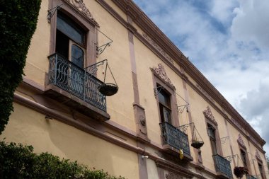 Balkonu ve sarı gökyüzü olan bir bina. Balkonda birkaç saksı bitkisi var. Queretaro şehrinin tarihi merkezi, Meksika 'nın Bağımsızlık Günü' nü, sömürge mimarisini, sokaklarını ve cephelerini kutlamak için dekorasyon ve gelenekler
