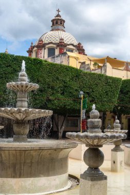 A fountain with a large dome on top of it. The fountain is surrounded by trees and has a green color. Historic center of Queretaro City, decorations and traditions to celebrate Mexico's Independence Day, colonial architecture, alleys and facades