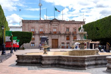 A large building with a fountain in front of it. The fountain is surrounded by trees. Historic center of Queretaro City, decorations and traditions to celebrate Mexico's Independence Day, colonial architecture, alleys and facades