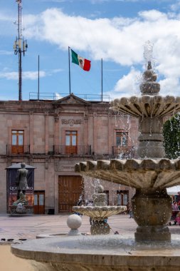 A large fountain with a flag flying above it. The flag is red, white, and green. Historic center of Queretaro City, decorations and traditions to celebrate Mexico's Independence Day, colonial architecture, alleys and facades