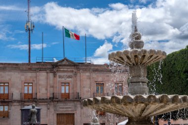 A large building with a fountain in front of it. A flag is flying in the background. Historic center of Queretaro City, decorations and traditions to celebrate Mexico's Independence Day, colonial architecture, alleys and facades