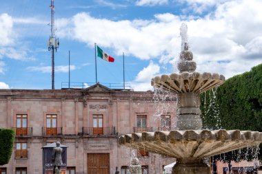 A large fountain with a flag in the background. The flag is red, white, and green. Historic center of Queretaro City, decorations and traditions to celebrate Mexico's Independence Day, colonial architecture, alleys and facades