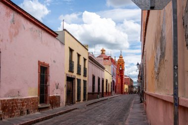 A street with a pink building and a red building. The street is narrow and has a cobblestone surface. Historic center of Queretaro City, decorations and traditions to celebrate Mexico's Independence Day, colonial architecture, alleys and facades
