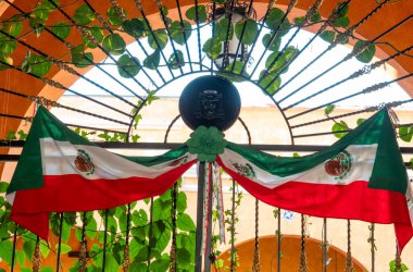 Büyük bir Meksika bayrağı dövülmüş demir bir kapının üzerine asılmış. Bayrak kırmızı, beyaz ve yeşildir. Queretaro şehrinin tarihi merkezi, Meksika 'nın Bağımsızlık Günü' nü, sömürge mimarisini, sokaklarını ve cephelerini kutlamak için dekorasyon ve gelenekler