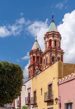 A building with two towers and a large tree in front of it. The building is yellow. The sky is blue. Historic center of Queretaro, colonial architecture, decorations for the celebration of Mexico's Independence Day.