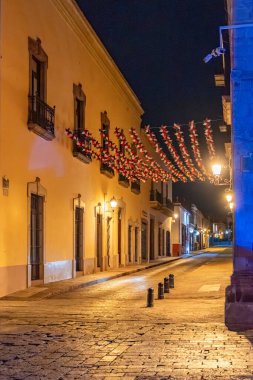 A street with a red and white banner hanging from a building. The banner is lit up at night. Historic center of Queretaro, colonial architecture, decorations for the celebration of Mexico's Independence Day.